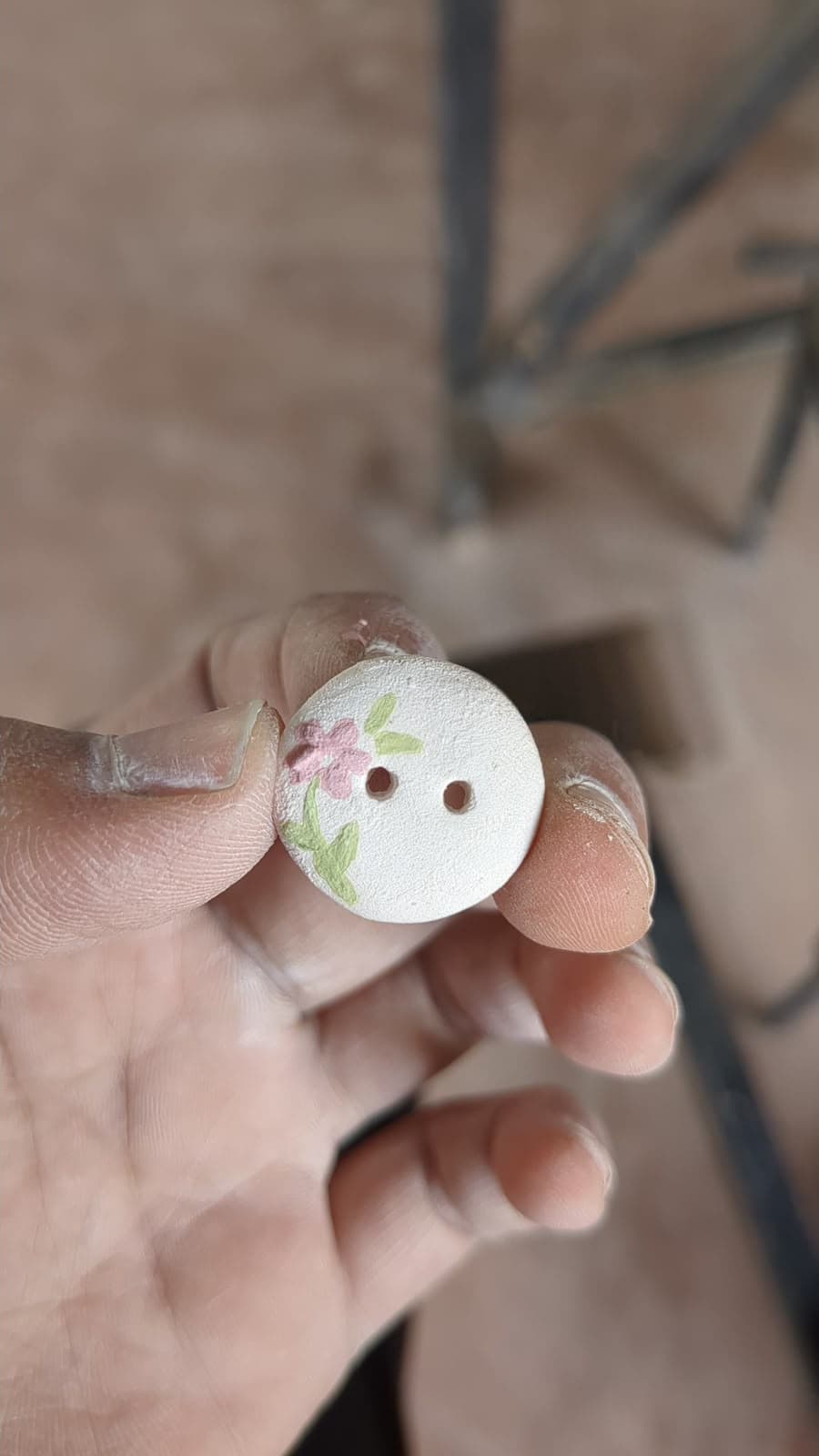 A hand-painted ceramic button, flowers on white, held between clay-dusted fingers