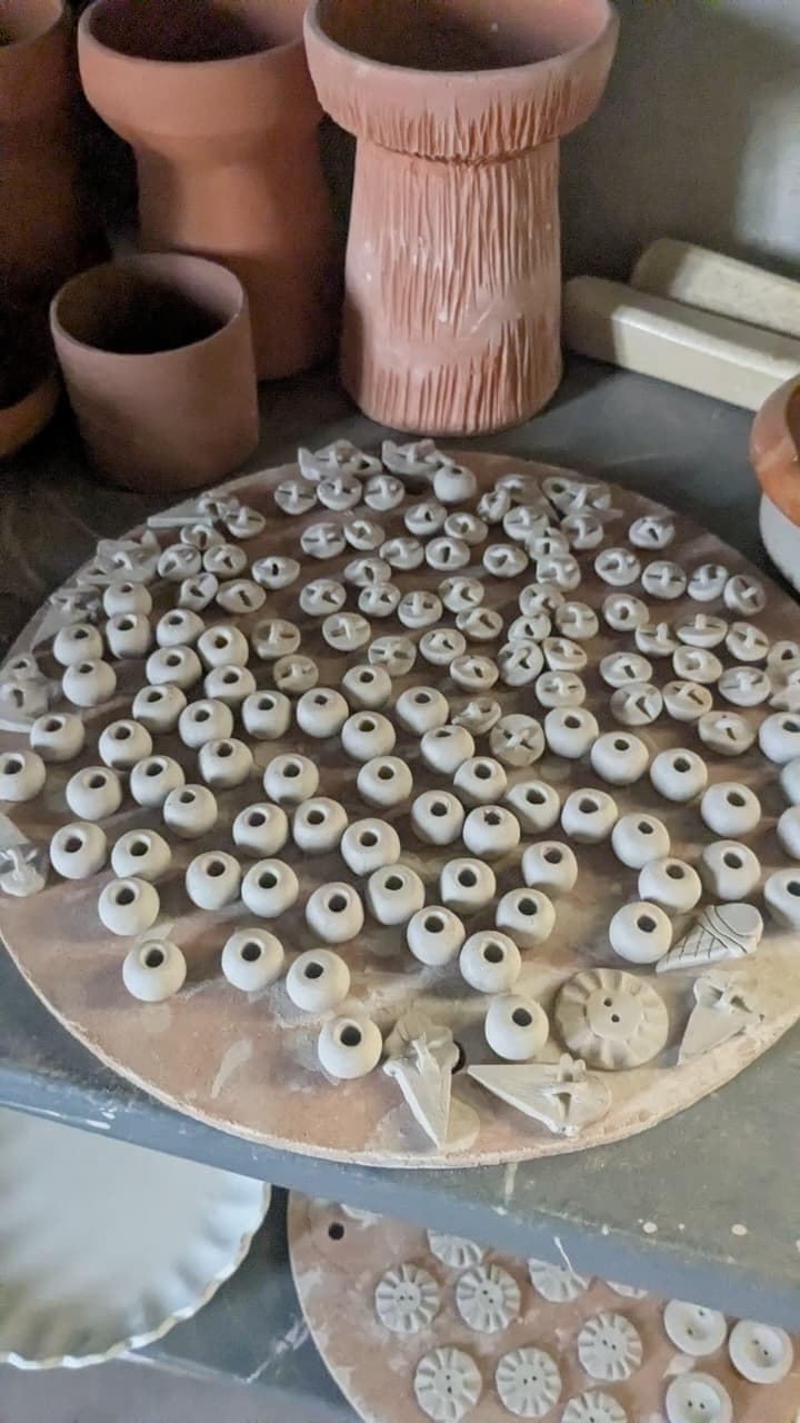 Unfired clay buttons and beads drying on a bat in the studio, thrown pots on the shelf behind